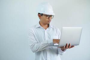 serious young asian architect man wearing builder safety helmet typing on the laptop isolated on white background photo