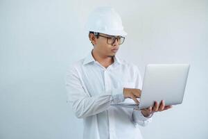 serious young asian architect man wearing builder safety helmet typing on the laptop isolated on white background photo