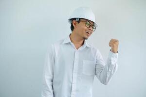 Young asian architect man wearing builder safety helmet over isolated background smiling with happy face looking and pointing to the side with thumb up. photo