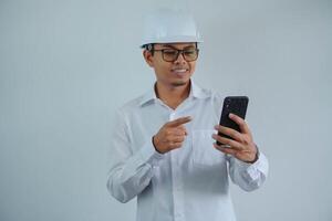 smiling young asian architect man wearing builder safety helmet pointing his mobile phone isolated on white background photo