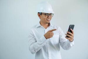 smiling young asian architect man wearing builder safety helmet pointing his mobile phone isolated on white background photo