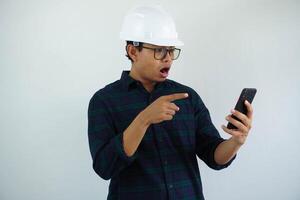 shocked young asian architect man wearing builder safety helmet pointing his mobile phone isolated on white background photo