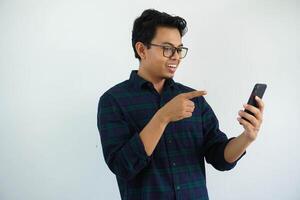 sonriente joven asiático hombre cuando mirando a su móvil teléfono y punto con un dedo aislado en blanco antecedentes foto