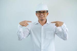 Young asian architect man wearing builder safety helmet smiling with happy face looking and pointing him self isolated on white background photo