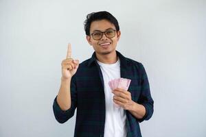 sonriente joven asiático hombre demostración contento cara expresión participación papel dinero mientras señalando su dedo arriba aislado en blanco antecedentes foto