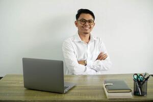 sonriente joven asiático empresario en un lugar de trabajo cruce su brazos con mirando cámara, vistiendo blanco camisa con lentes aislado foto