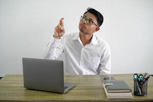 young asian businessman in a workplace with fingers crossing and wishing the best wearing white shirt isolated on white background photo