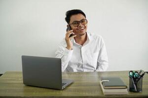 smiling young asian businessman in a workplace keeping a conversation with the mobile phone wearing white shirt with glasses isolated photo
