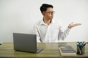 young asian businessman in a workplace holding copy space with doubts, wear white shirt with glasses isolated photo