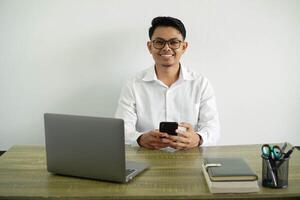 young asian businessman in a workplace smiling sending a message with the mobile phone, wearing white shirt with glasses isolated photo