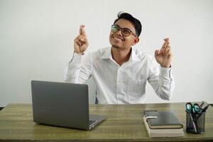 young asian businessman in a workplace with fingers crossing and wishing the best, wear white shirt with glasses isolated photo