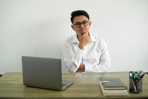 young asian businessman in a workplace thinking with holding chin, wear white shirt with glasses isolated photo