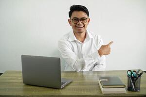 young asian businessman in a workplace smiling with pointing back, wearing white shirt with glasses isolated photo