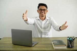 young asian businessman in a workplace smiling with both arms open to welcome and inviting to come with hand, wearing white shirt with glasses isolated photo