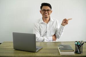 smiling young asian businessman in a workplace pointing finger to the side, wearing white shirt with glasses isolated photo