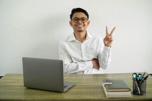 joven asiático empresario en un lugar de trabajo sonriente y demostración victoria firmar, vistiendo blanco camisa con lentes aislado foto