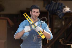 Portrait of a carpenter holding a spirit level and handsaw in his workshop. photo