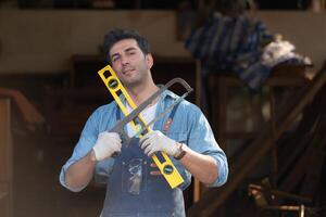 Portrait of a carpenter holding a spirit level and handsaw in his workshop. photo