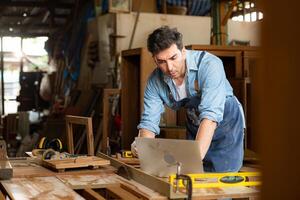 Carpenter working on laptop in his workshop at a woodworking factory photo