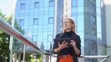 joven mujer de negocios participación tableta en pie en ciudad calle. hermosa rubia mirando a artilugio pantalla, en verano día. concepto. negocio video