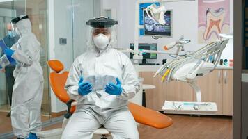 Dentistry doctor wearing protection suit speaking on camera with patients during pandemic coronavirus. Orthodontic on video call with coverall, face shield, mask, gloves with assistant in background photo