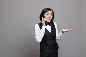 Catering service employee in uniform explaining to customer while answering call on smartphone. Young asian woman receptionist having conversation with hotel staff on mobile phone photo