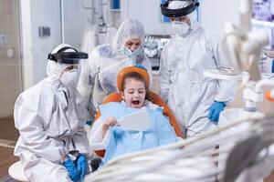 Little girl smiling looking in mirror sitting on dental chair after oral hygine procedure dressed in ppe suit. Child wearing ppe suit during teeth intervention at dental hospital. photo