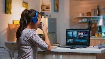 Woman video editor with headset working with footage and sound sitting in home kitchen. Woman videographer editing audio film montage on professional laptop sitting on desk in midnight photo