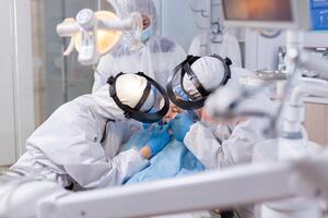 Orthodentist and assistant treating little girl using modern instruments dressed in ppe suit. Stomatology team wearing ppe suit during covid19 doing procedure on child teeth. photo