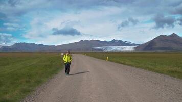 Female tourist with backpack goes by dirt road on background of mountains and glacier in Iceland. Freedom and travel concept. 4K video