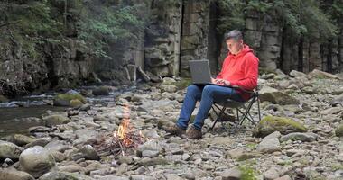 een Mens werken Aan een laptop in de buurt een stroom in natuur. concept van freelancen, digitaal nomade of afgelegen kantoor. 4k video