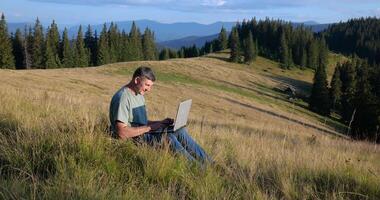 ein Mann sitzt auf ein schön Wiese im das Berge, funktioniert auf ein Laptop. Konzept von freiberuflich, Digital Nomade oder Fernbedienung Büro. video