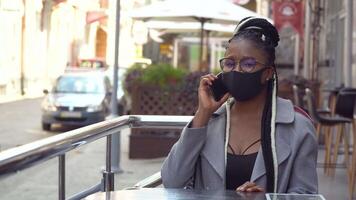 Young black woman in mask talks on phone in street cafe. Beautiful female using mobile phone to talk video