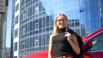 atractivo mujer de negocios con largo pelo cerca lujo coche mirando a el cámara en un edificio antecedentes video