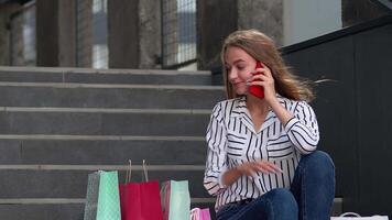 Girl sitting on stairs with bags talking on cell phone about sale in shopping mall in Black Friday video