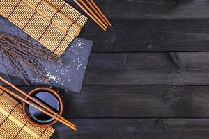 Background for sushi. Bamboo mat and soy sauce on black wooden table. Top view with copy space photo