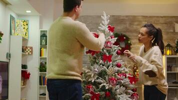 Young couple in love assembled christmas tree in living room. Romantic couple. Decorating beautiful xmas tree with glass ball decorations. Wife and husband in matching clothes helping ornate home with garland lights video