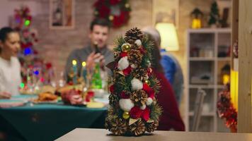 weinig Kerstmis boom Aan focus met familie vieren Kerstmis in de achtergrond. traditioneel feestelijk Kerstmis avondeten in multigenerationeel familie. genieten van Kerstmis maaltijd feest in versierd kamer. groot familie bijeenkomst video