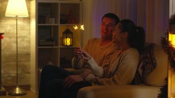 vrolijk man en zijn vrouw spelen met sparkles Aan Kerstmis dag. paar kussen. vieren nieuw jaar vooravond en Kerstmis nacht, samen liefde viering sterretje en haard in achtergrond video