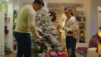 caucasian man och kvinna Lycklig till dekorera deras jul träd. vinter- högtider. dekorera skön xmas träd med glas boll dekorationer. fru och Make i matchande kläder portion utsmyckad Hem med krans lampor video