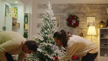 alegre joven Pareja Decorar Navidad árbol y esperando para Papa Noel. decorando hermosa Navidad árbol con vaso pelota decoraciones esposa y marido en pareo ropa Ayudar florido hogar con guirnalda luces video