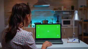 Woman looking at laptop with green mockup during night time in home kitchen. Freelancer watching desktop monitor display with green screen, chroma key, during night time working overtime. photo