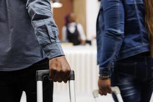 Close up of African American couple arriving at hotel or resort, tourists standing with suitcases in lobby and waiting for check-in process. Cropped photo of man and woman pulling luggage