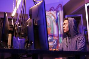 Asian hacker using spyware to access database on computer in abandoned warehouse with graffiti. Young man in hood breaking law while programming illegal malicious malware photo