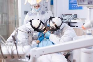 Little girl during dentist visit getting oral hygine treatment from dentistiry team. Stomatology team wearing ppe suit during covid19 doing procedure on child teeth. photo