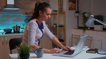 Woman checking watch during overtime work in home kitchen at midnight closing laptop and leaving. Busy focused employee using modern technology network wireless reading typing, searching photo