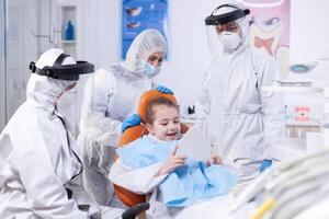 Excited kid dresse cover all holding mirror after stomatology procedure sitting on chair. Child wearing ppe suit during teeth intervention at dental hospital. photo