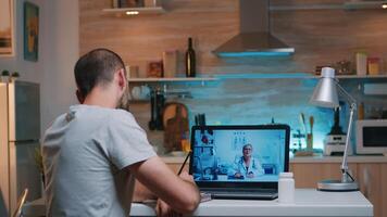 Male writing on notebook during online consultation listening woman doctor sitting in front of laptop in the kitchen. Sick man discussing during video conference about symptoms holding bottle of pills photo