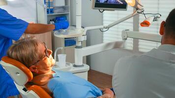 Old woman at dentist having dental treatment in implantology office. Orthodontist lighting the lamp and speaking to patient sitting on stomatological chair while nurse preparing tools for surgery. photo