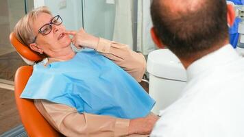 cerca arriba de mayor mujer quejumbroso a estomatólogo acerca de doloroso sentimientos en dientes. mayor paciente explicando dental problema a odontología médico indicando boca mientras examinando radiografía. foto